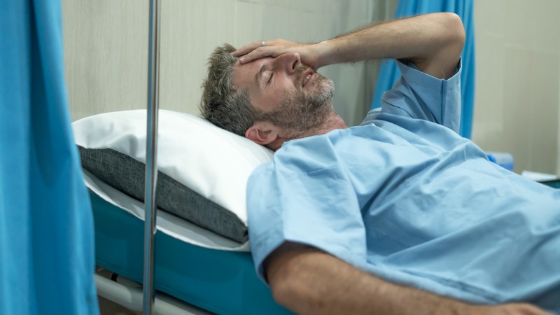 A male patient looks pained and frustrated while lying in a hospital bed.