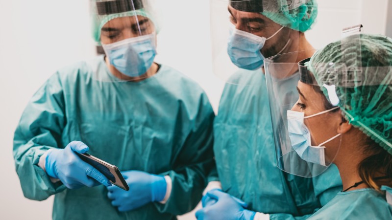 Three physicians wearing scrubs masks, and face shields consult. One is holding up a smartphone that the others can see.