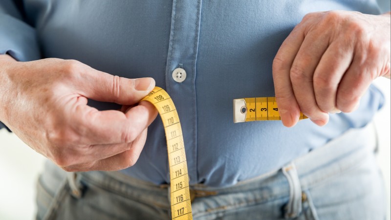 A man measures his waistline using a yellow tape measure.